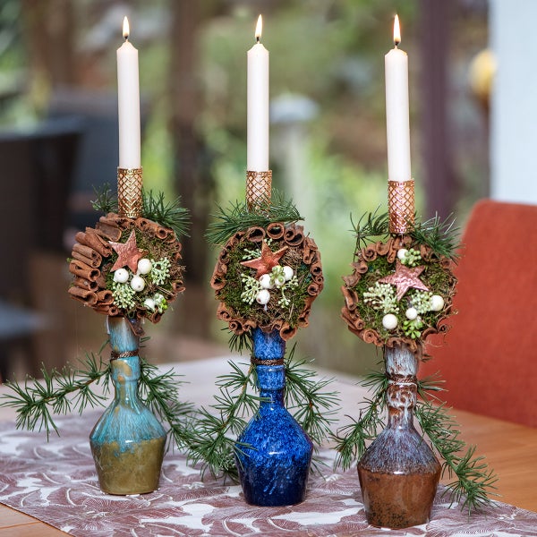 Drei festlich dekorierte Kerzenhalter mit weißen Stabkerzen, verziert mit Zimtstangen, Moos, Beeren und kleinen Sternen.