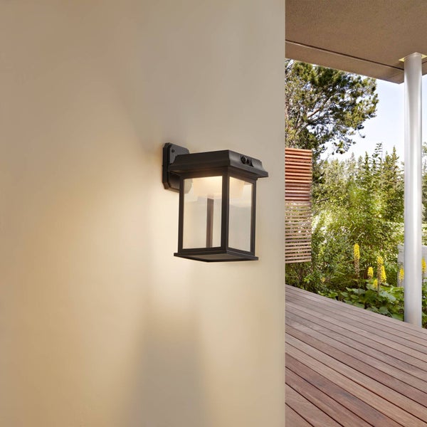 Schwarze Außenwandleuchte im Laternendesign mit Glaseinsätzen, beleuchtet an einer Wand montiert neben einer Holzterrasse und Garten.