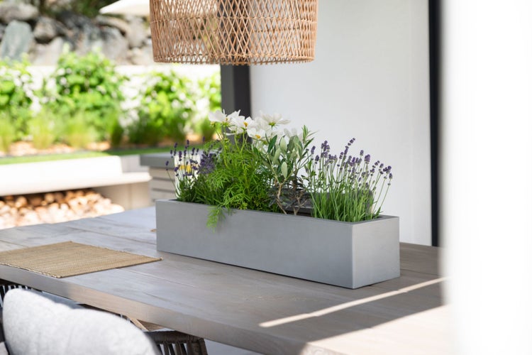 Moderner grauer rechteckiger Blumenkasten mit Lavendel und weißen Blumen auf einem hölzernen Gartentisch unter einer Korblampe.