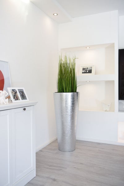 Hohe, silberfarbene Bodenvase mit künstlichem Gras in einem hellen, modern eingerichteten Wohnzimmer neben einer weißen Kommode.