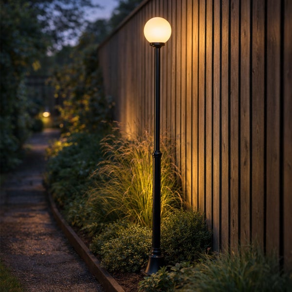 Schwarze Außenstandleuchte mit leuchtender Kugelform in einem Gartenbeet neben einem Holzzaun und einem gepflasterten Weg bei Dämmerung.