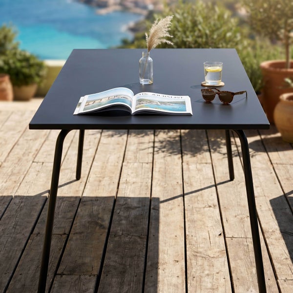 Rechteckiger dunkelgrauer Gartentisch auf einer Holzterrasse mit Blick auf das Meer, dekoriert mit einer Zeitschrift und einem Glas.