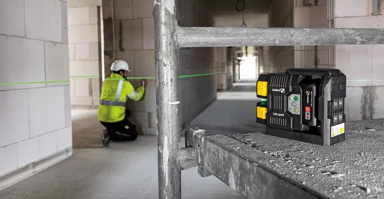 Ein Stabila Kreuzlinienlaser LAX 500G auf einem Baugerüst, während ein Handwerker im Hintergrund eine grüne Laserlinie an einer Wand markiert.