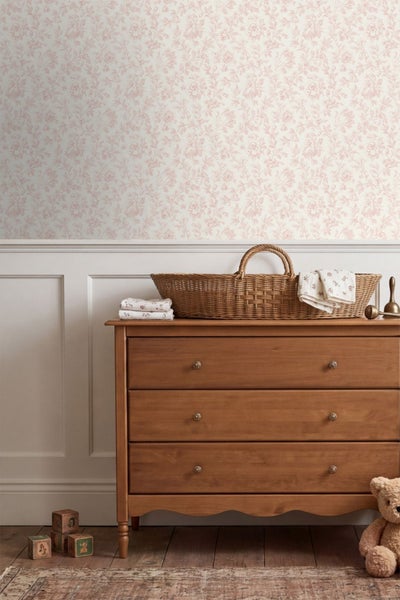 Vliestapete mit floralem Muster über weißen Wandpaneelen in einem hellen Kinderzimmer mit Holzkommode und Flechtkorb.