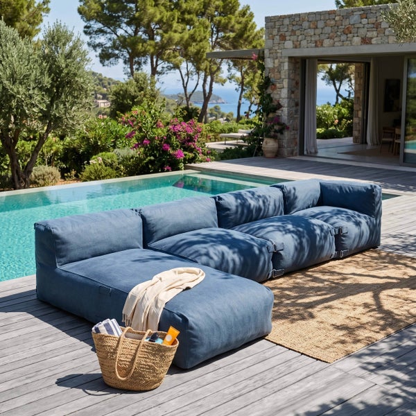 Modulare blaue Outdoor-Gartenlounge auf einer Holzterrasse neben einem Swimmingpool mit Garten und Steinhaus im Hintergrund.