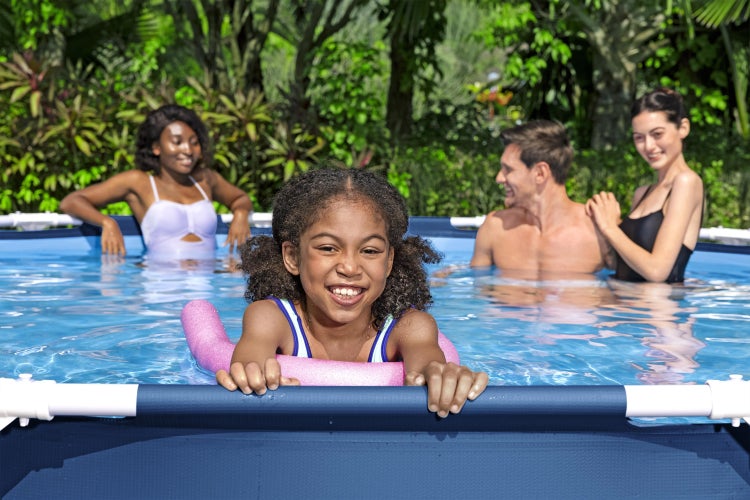 Familie in einem hellen Aufstellpool mit Metallrahmen im Garten. Ein Kind nutzt eine Poolnudel als Schwimmhilfe am Beckenrand.