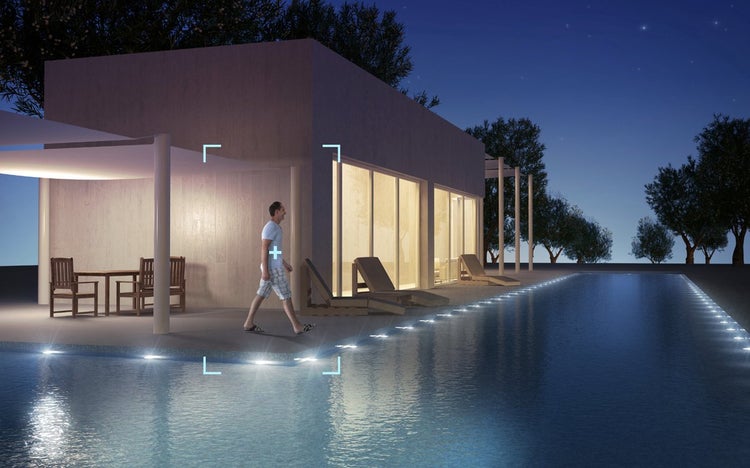 Moderne Terrasse mit Pool bei Nacht, beleuchtet durch Bodeneinbaustrahler am Beckenrand. Eine Person geht an Liegestühlen und einem Tisch vorbei.