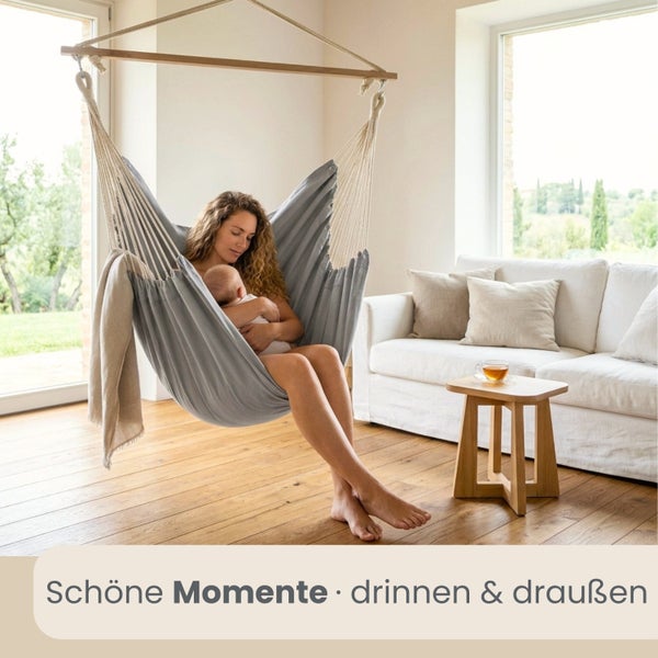 Eine Frau mit Baby in einem grauen Hängesessel in einem hellen Wohnzimmer mit Parkettboden, weißem Sofa und Holzbeistelltisch.