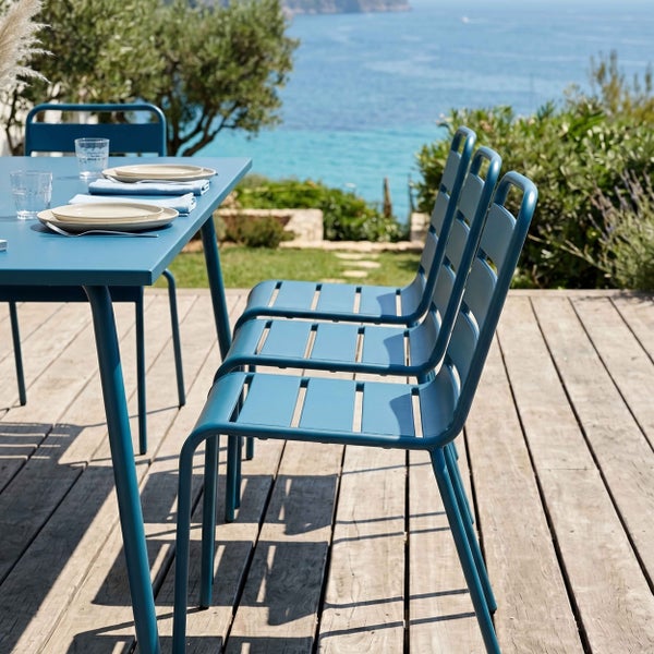 Gartenmöbel-Set aus blauem Metall auf einer Holzterrasse mit Meerblick, bestehend aus einem Tisch und Lamellenstühlen.