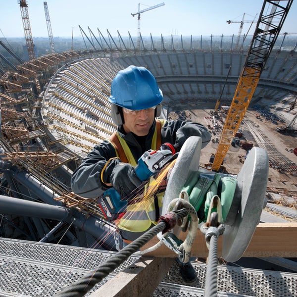 Bauarbeiter mit Schutzausrüstung nutzt einen Akkumulator Winkelschleifer auf einer Stadion Baustelle mit Kränen im Hintergrund.