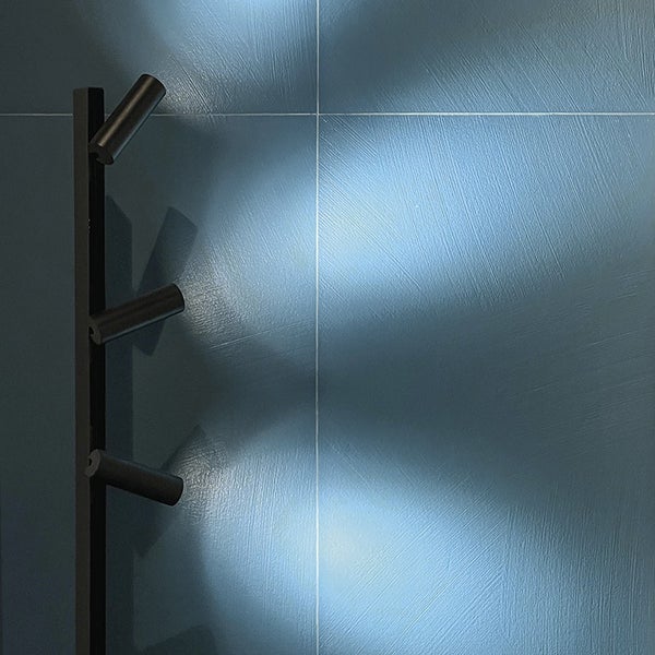 Wandleuchte mit drei Strahlern an einer vertikalen Schiene, die Lichtkegel auf eine strukturierte Wandfliese wirft.