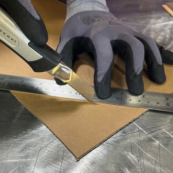 Messer schneidet Pappe entlang eines Metalllineals, getragen werden Arbeitshandschuhe