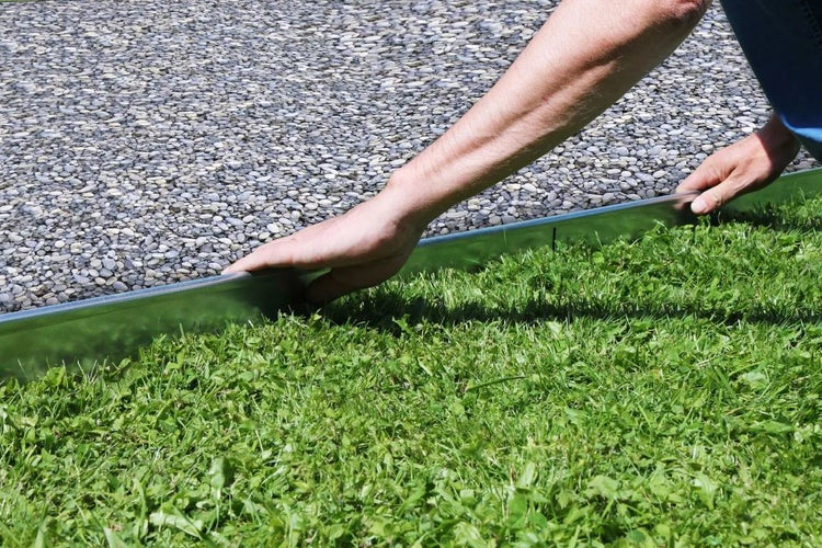 Eine Person verlegt eine Rasenkante aus Metall zwischen Rasen und Kies.