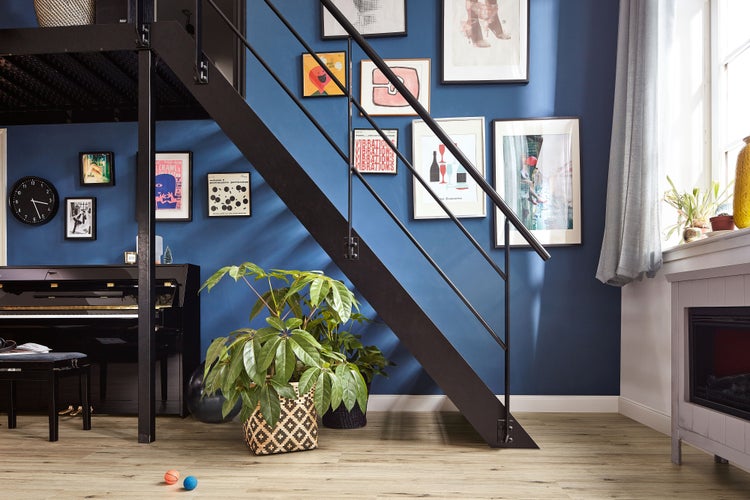 Wohnraum mit blauen Wänden, schwarzer Metalltreppe, Klavier, Wandgalerie mit gerahmten Bildern und Zimmerpflanzen auf einem Holzboden.