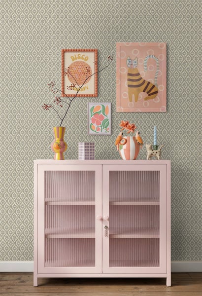 Vliestapete mit dezentem geometrischem Muster an einer Wand hinter einer rosa Metallkommode mit geriffelten Glastüren und farbenfroher Dekoration.
