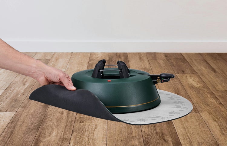 Hand hebt graue Schutzunterlage mit Rentier- und Schneeflockenmuster unter grünem Christbaumständer auf Holzboden an.