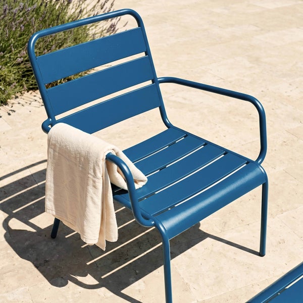 Blauer Gartenstuhl aus Metall mit Armlehnen und Lattenstruktur auf einer hellen Terrasse, ein Handtuch hängt über der Armlehne.