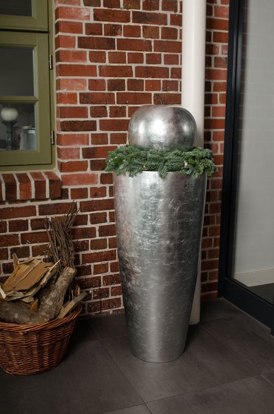 Silberne Bodenvase mit Tannenkranz und dekorativem Apfel vor einer Ziegelwand neben einem Korb mit Kaminholz.