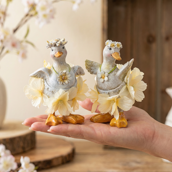 Zwei dekorative Entenfiguren mit Blumenkranz und Blütenrock, auf einer Hand präsentiert.