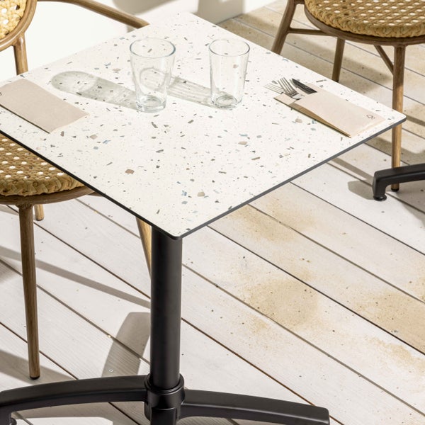 Quadratischer Bistrotisch mit Platte in Terrazzo-Optik und schwarzem Metallfuß auf einer Holzterrasse mit Gläsern und Besteck.