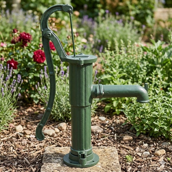 Handbetriebene Schwengelpumpe aus Gusseisen auf einem Steinsockel in einem bepflanzten Gartenbeet.