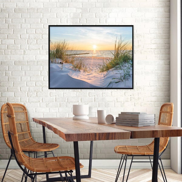 Wandbild mit Strandmotiv in schwarzem Rahmen über einem Holztisch mit Rattan-Stühlen vor einer hellen Backsteinwand.