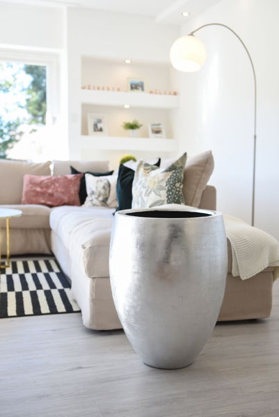Großer silberner Pflanzkübel in einem hellen, modernen Wohnzimmer mit Sofa und gestreiftem Teppich.