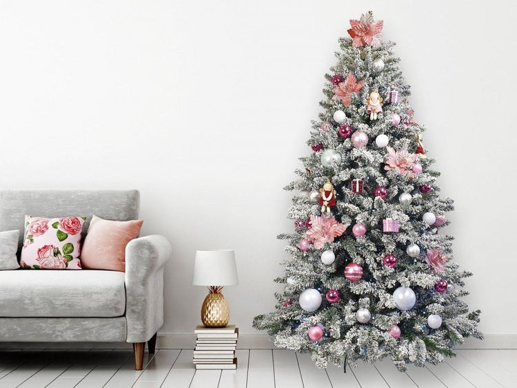 Ein geschmückter, künstlicher Weihnachtsbaum mit Schneeoptik, dekoriert mit Kugeln und Blüten, neben einem Sofa und einer kleinen Tischleuchte.