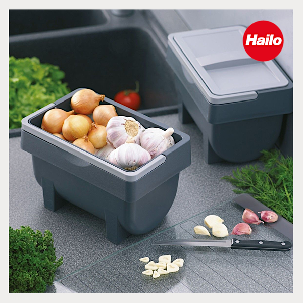 Hailo Logo. Graue Küchen-Aufbewahrungsboxen auf einer Arbeitsplatte neben einer Spüle, befüllt mit Zwiebeln und Knoblauch.