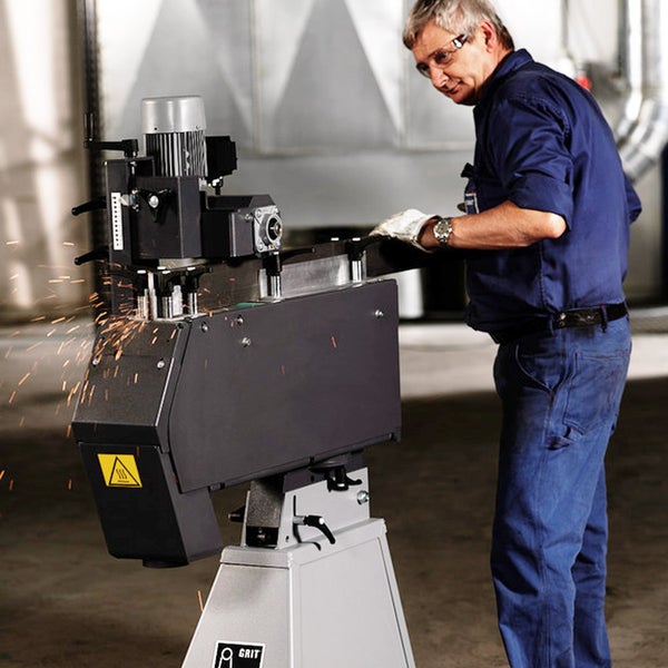 Ein Mann bedient eine stationäre Metallschleifmaschine der Marke GRIT, beim Schleifen eines Werkstücks entstehen Funken.