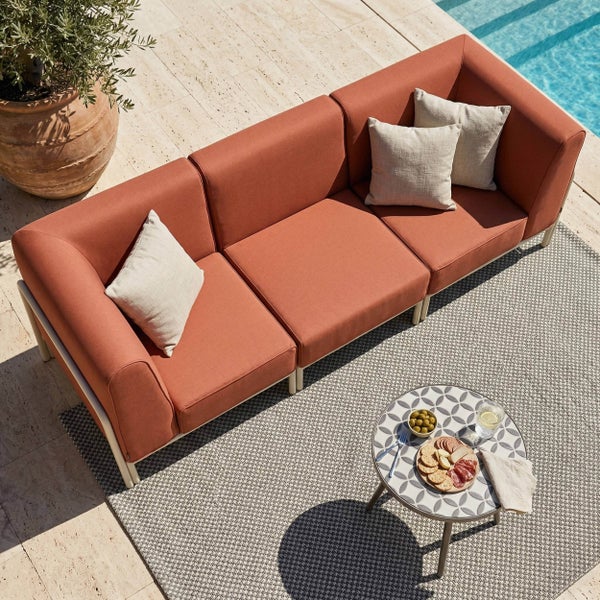 Modernes 3-Sitzer-Outdoor-Sofa mit rostfarbenen Polstern, runder Beistelltisch und Außenteppich auf einer Steinterrasse am Pool.