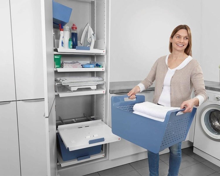 Frau hält Wäschekorb neben organisiertem Hochschrank mit Auszügen für Bügeleisen und Waschmittel in einem hellen Hausarbeitsraum.