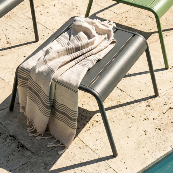 Anthrazitfarbener Fußhocker aus Aluminium auf einer hellen Steinterrasse am Pool mit gestreiftem Baumwolltuch in moderner Außenumgebung.