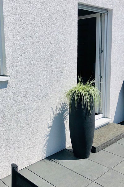 Hoher, schwarzer Pflanzkübel mit Ziergras auf einer modernen Terrasse neben einer weißen Hauswand.