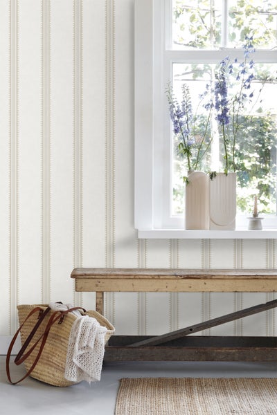Wohnraum mit Vliestapete mit Streifenmuster, Holzbank am Fenster mit Blumen in Vasen und Korbtasche.