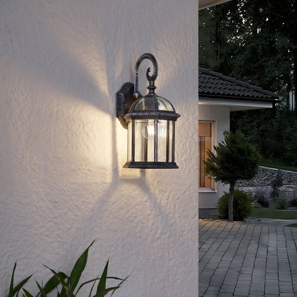 Klassische Außenwandleuchte im Laternen-Design an einer weißen strukturierten Wand, beleuchtet mit warmem Licht, Außenbereich mit Terrasse und Garten.