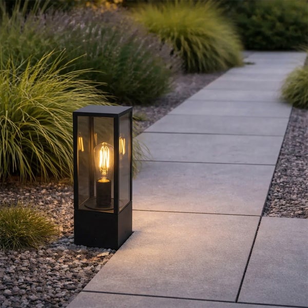 Moderne schwarze Sockelleuchte mit Glaseinsatz und leuchtender Filamentlampe an einem Gartenweg aus Steinplatten und Ziergräsern.