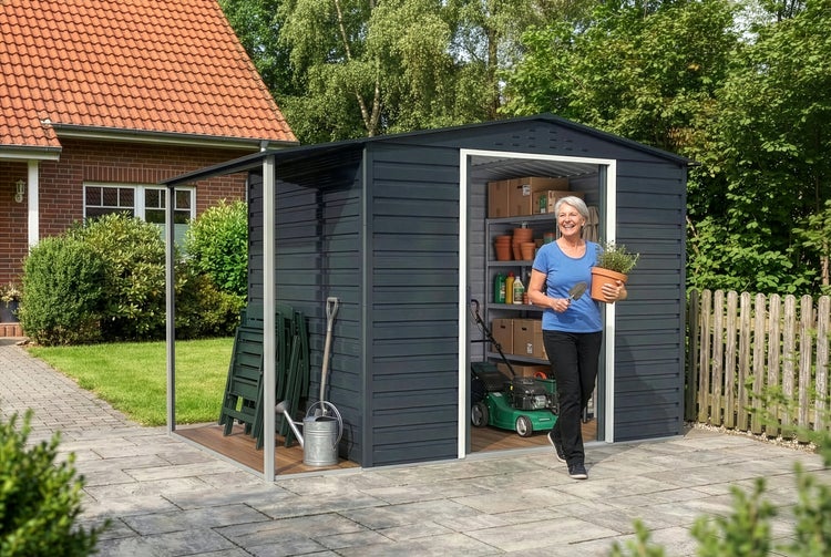 Dunkelgraues Metallgerätehaus mit Schleppdach auf heller Terrasse, Frau mit Topfpflanze tritt heraus, Innenraum mit Regalen und Rasenmäher.