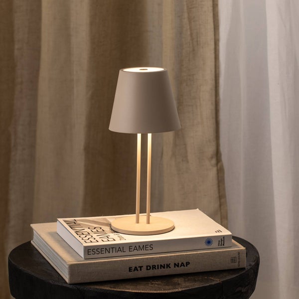 Moderne Tischleuchte mit konischem Schirm und schlankem Doppelstabstativ auf Büchern, warm beleuchtet vor Vorhängen.