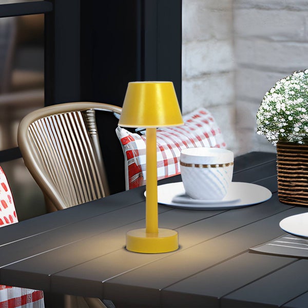 Moderne gelbe LED-Tischleuchte auf einem dunklen Gartentisch mit Stühlen und Geschirr im Hintergrund.
