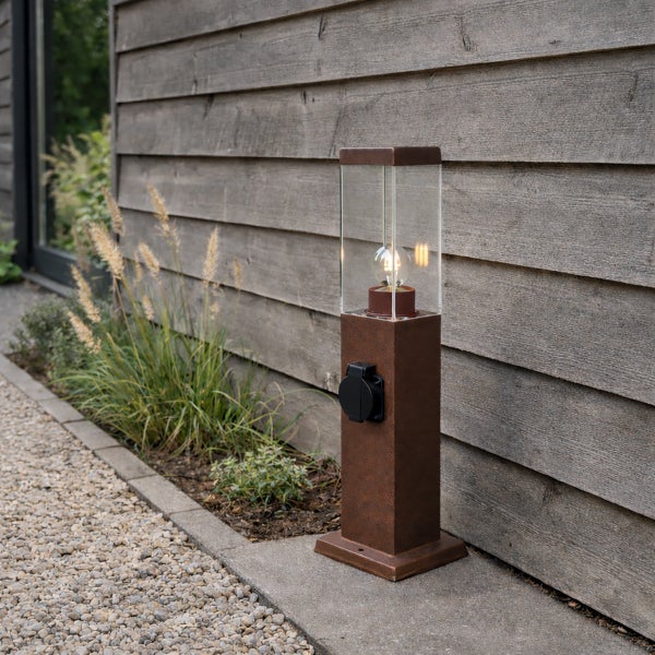 Rustikale Außenleuchte in Rostoptik mit integrierter Steckdose und Glaseinsatz vor einer Holzfassade im Garten.