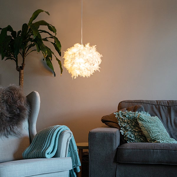 Beleuchtete Feder-Pendelleuchte in einem gemütlichen Wohnzimmer mit grauem Sofa, Sessel und Zimmerpflanze.