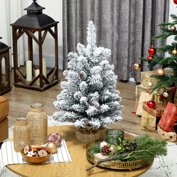 Kleiner, künstlicher Weihnachtsbaum, beschneit, mit Zapfen auf einem dekorierten Tisch in einem hellen Wohnzimmer mit weiterer Weihnachtsdekoration.