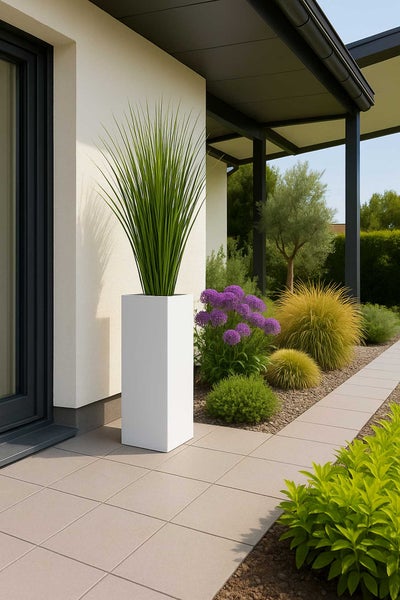 Hoher quadratischer weißer Pflanzkübel mit Ziergras auf einer modernen Fliesenterrasse neben einem Gartenbeet.
