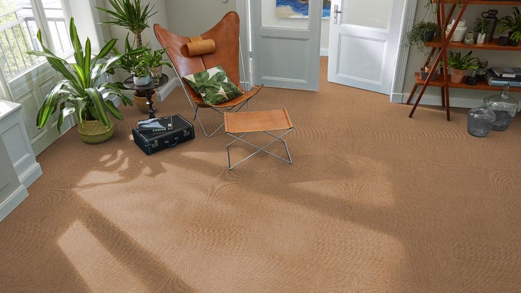 Moderner Wohnraum mit braunen, strukturierten Teppichfliesen, ledernem Butterfly-Sessel, Zimmerpflanzen und hellem Lichteinfall.