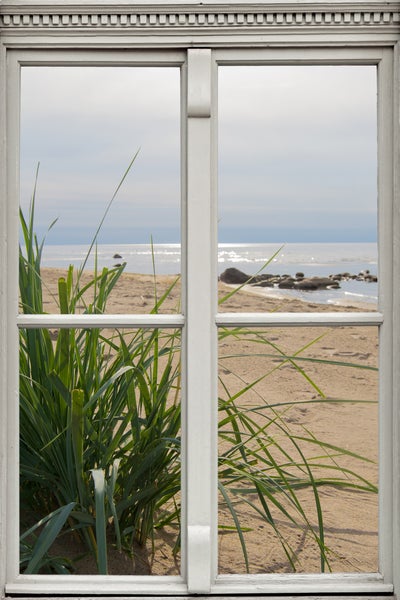 Blick durch ein Fenster auf einen Strand mit Gras und Meer