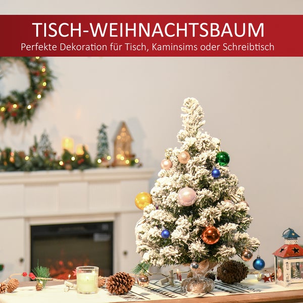 Beschneiter Tisch-Weihnachtsbaum mit bunten Kugeln und Kerze auf einem festlich dekorierten Tisch vor einem Kamin.