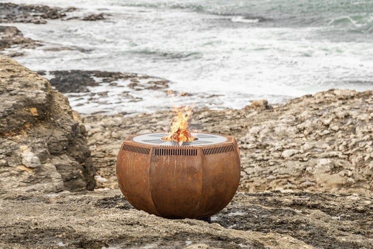 Feuerschale in Kugelform aus Rostoptik Metall mit loderndem Feuer in felsiger Küstenlandschaft am Meer