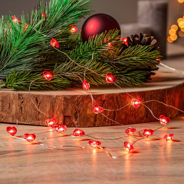 LED-Lichterkette mit herzförmigen Lichtern an silberfarbenem Draht, dekoriert auf einer Holzscheibe mit Tannenzweigen und einer Kugel.