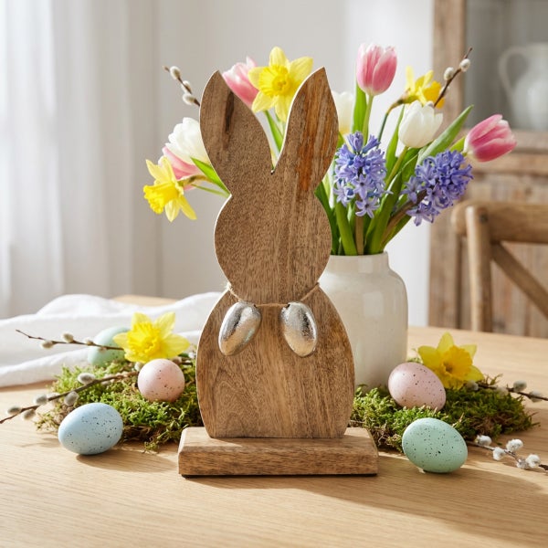Osterdekoration aus Holz in Hasenform mit zwei kleinen Eiern an einer Schnur, platziert auf einem Holztisch mit Blumenstrauß und Ostereiern.